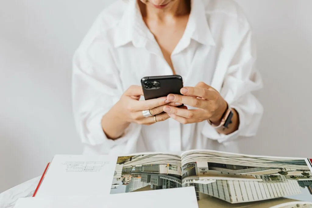 A woman wearing a white shirt on her smart phone with magazines open in front of her