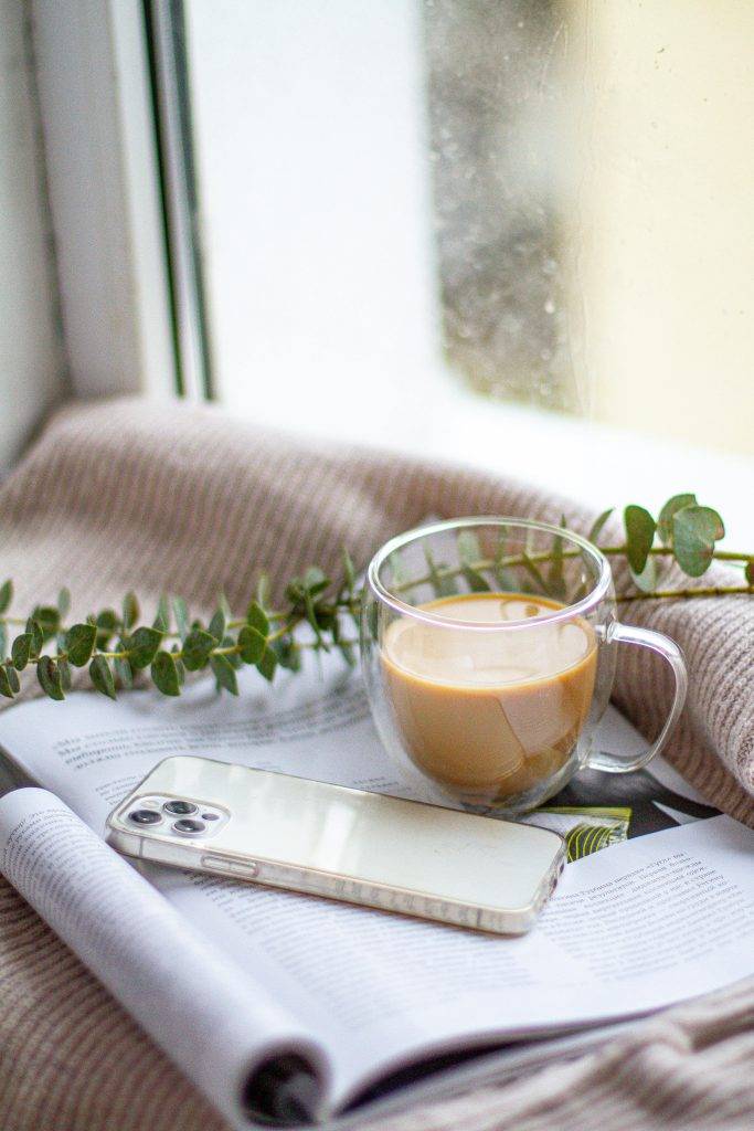 A mobile phone resting on an open magazine with a hot drink and some greenery