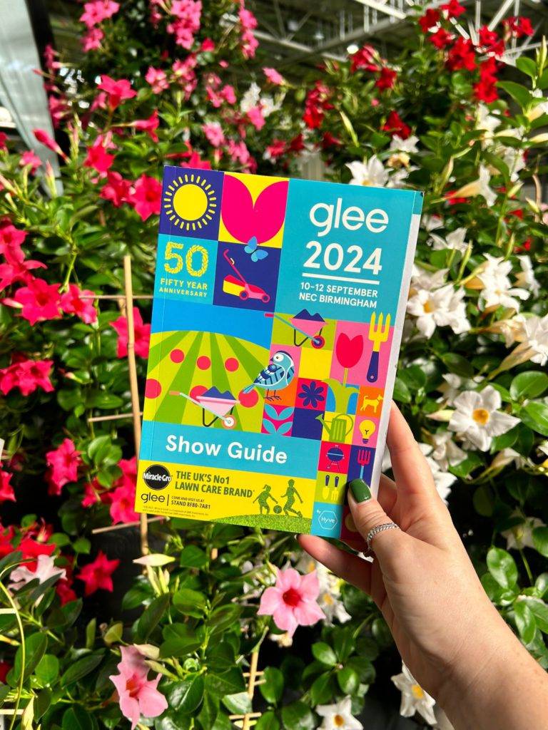 The Glee 2024 exhibition programme being held up against a wall of flowers