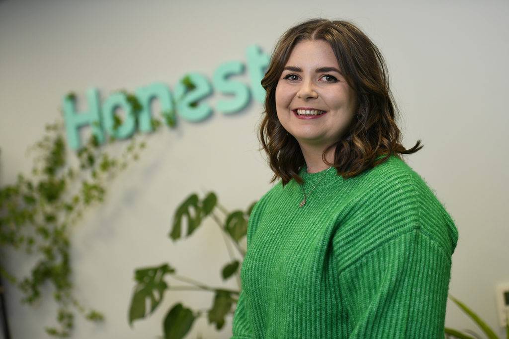 A woman wearing a green jumper stood in front of an "honest" sign smiling at the camera